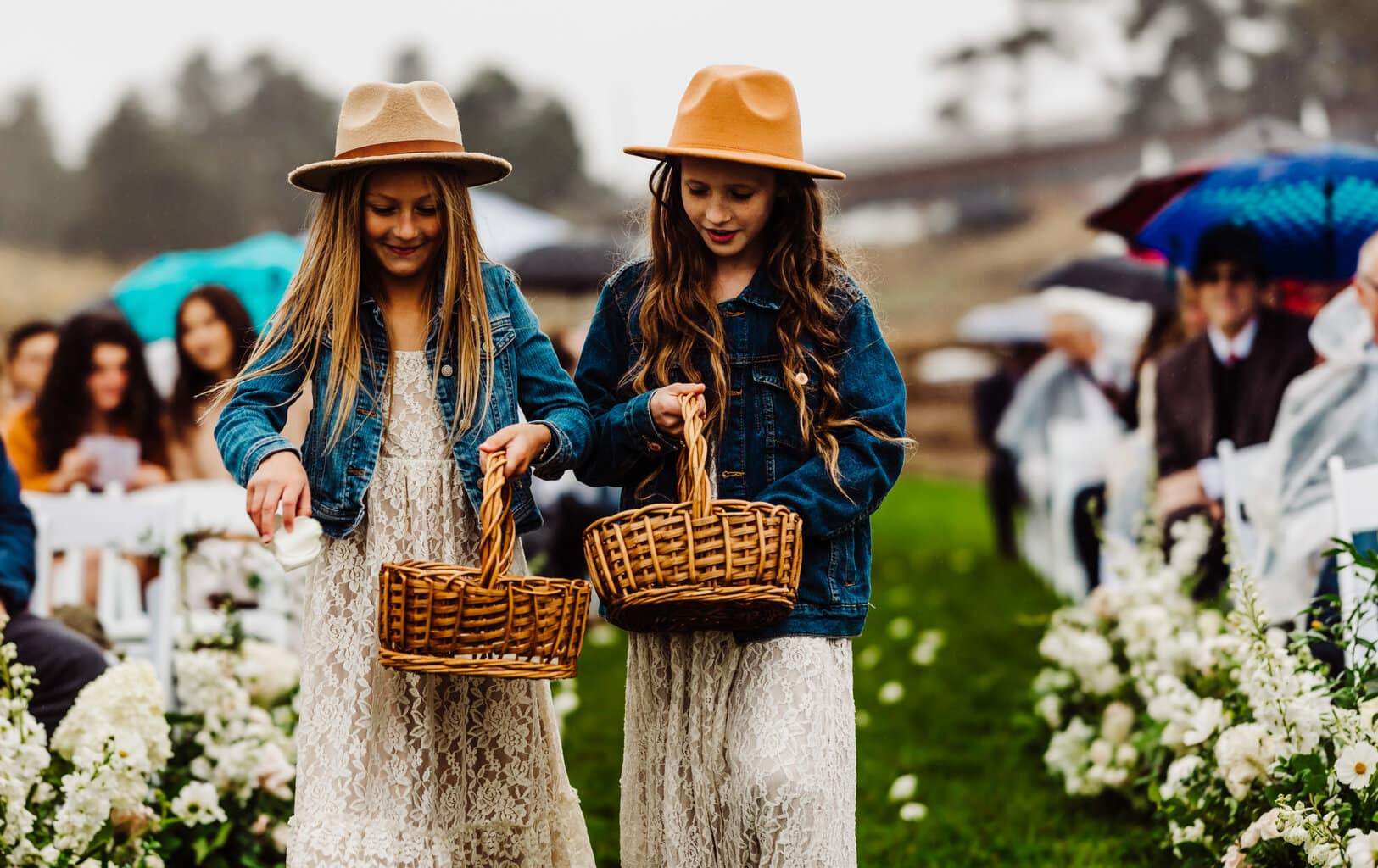 Colorado Wedding Photographer | The photograph shows two young flower girls walking down the aisle at a wedding. They are holding wicker baskets that are filled with flowers and throwing the flowers on the outdoor aisle as they walk towards the altar. The girls are dressed in lacy white, bohemian style dresses. They have on jean jackets and tan brimmed hats.