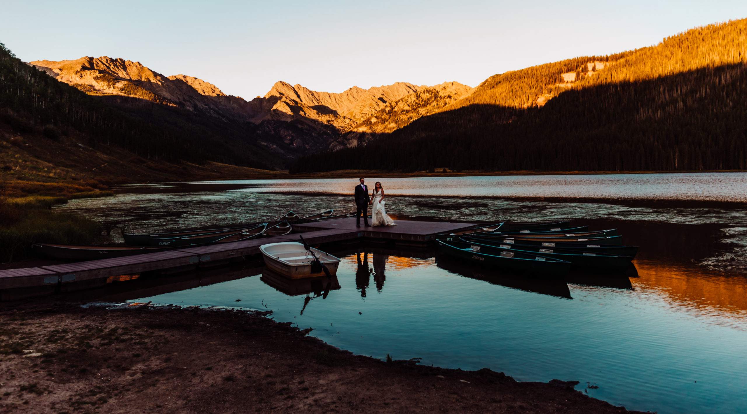 Colorado Wedding Photographer | The photograph is of a couple at their summer wedding at Piney River Ranch outside of Vail, Colorado. The couple is standing on a dock at sunset looking at the camera smiling. The background has beautiful mountain scape with alpenglow hitting the mountains perfectly.